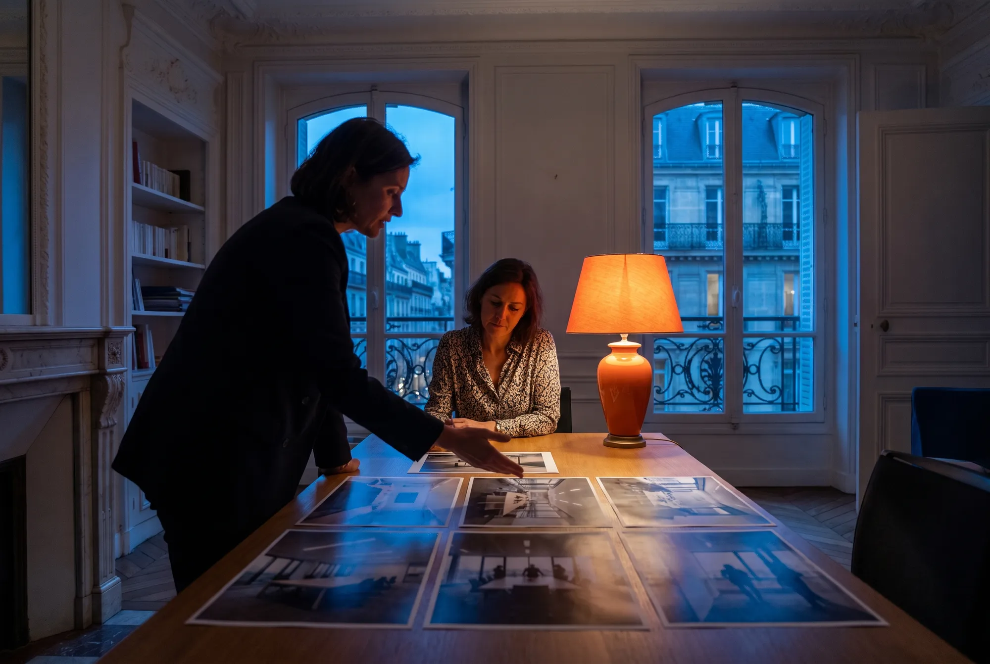 Productrice présentant des photos de repérage imprimées à une responsable communication dans un bureau haussmannien le soir