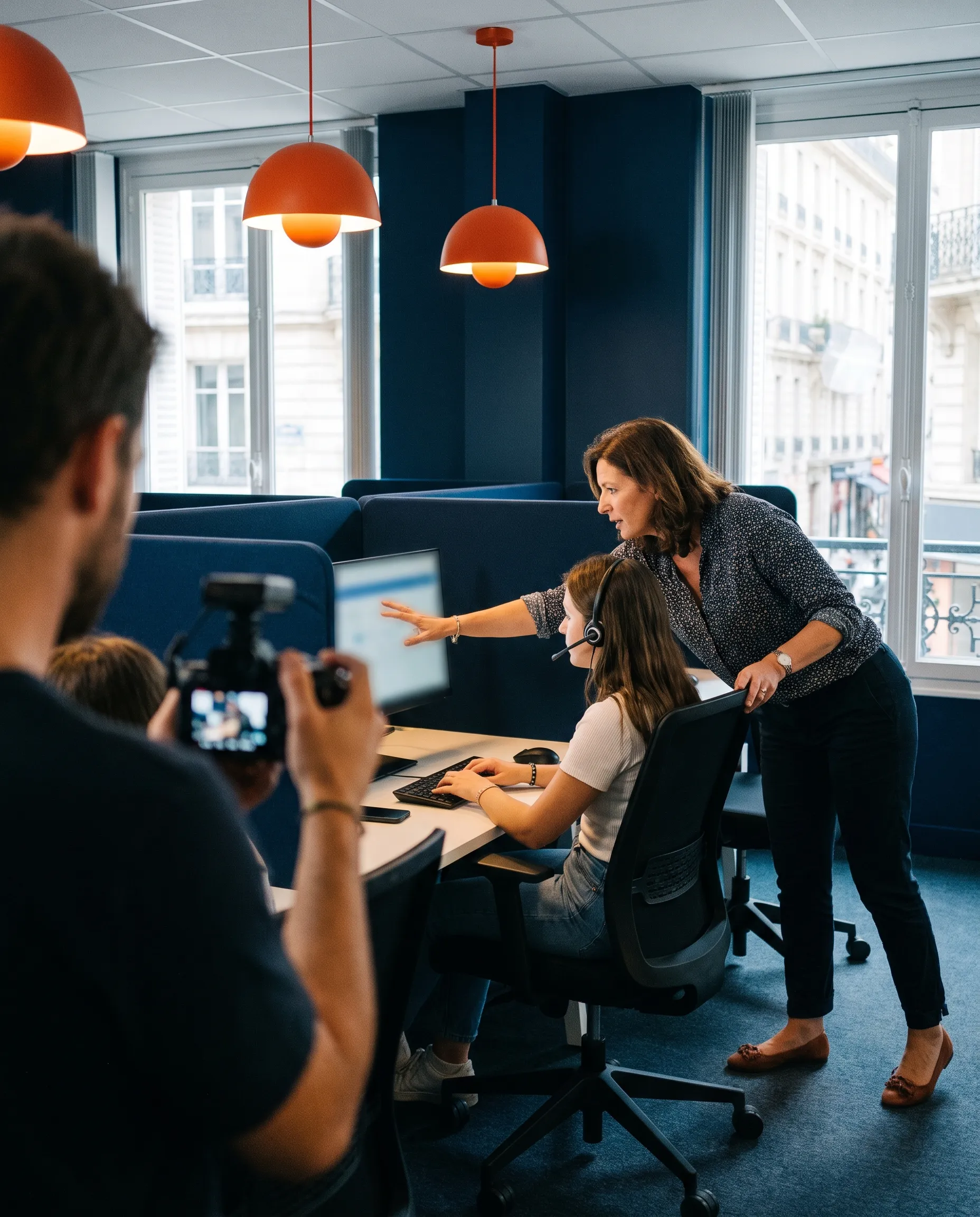 Formatrice interne guidant une conseillère équipée d’un casque-micro sur son poste, filmée par un vidéaste dans le centre de relation client d’une entreprise