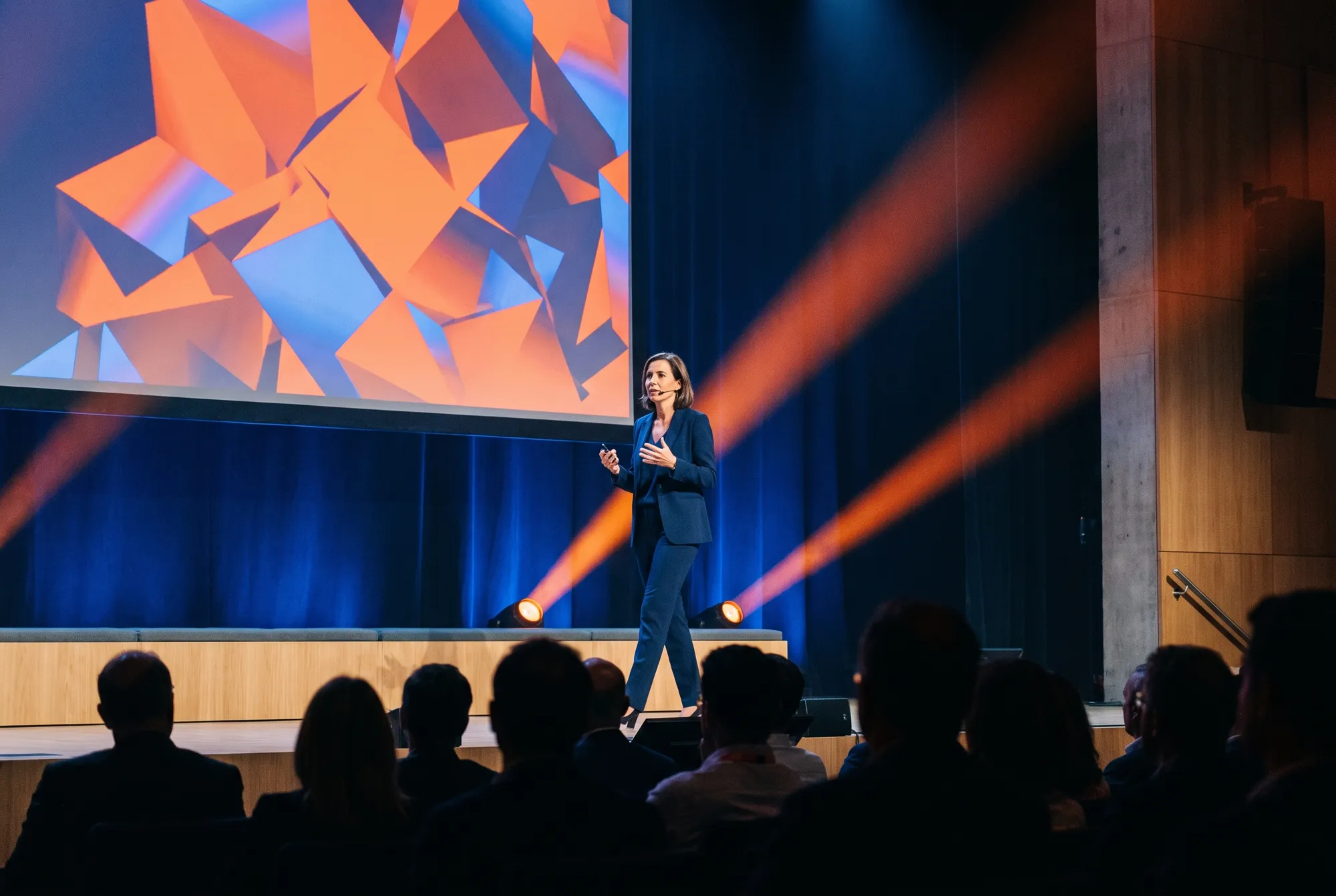 Intervenante en tailleur bleu nuit s’adressant au public sur la scène d’un auditorium, devant un grand écran de formes orange et bleues