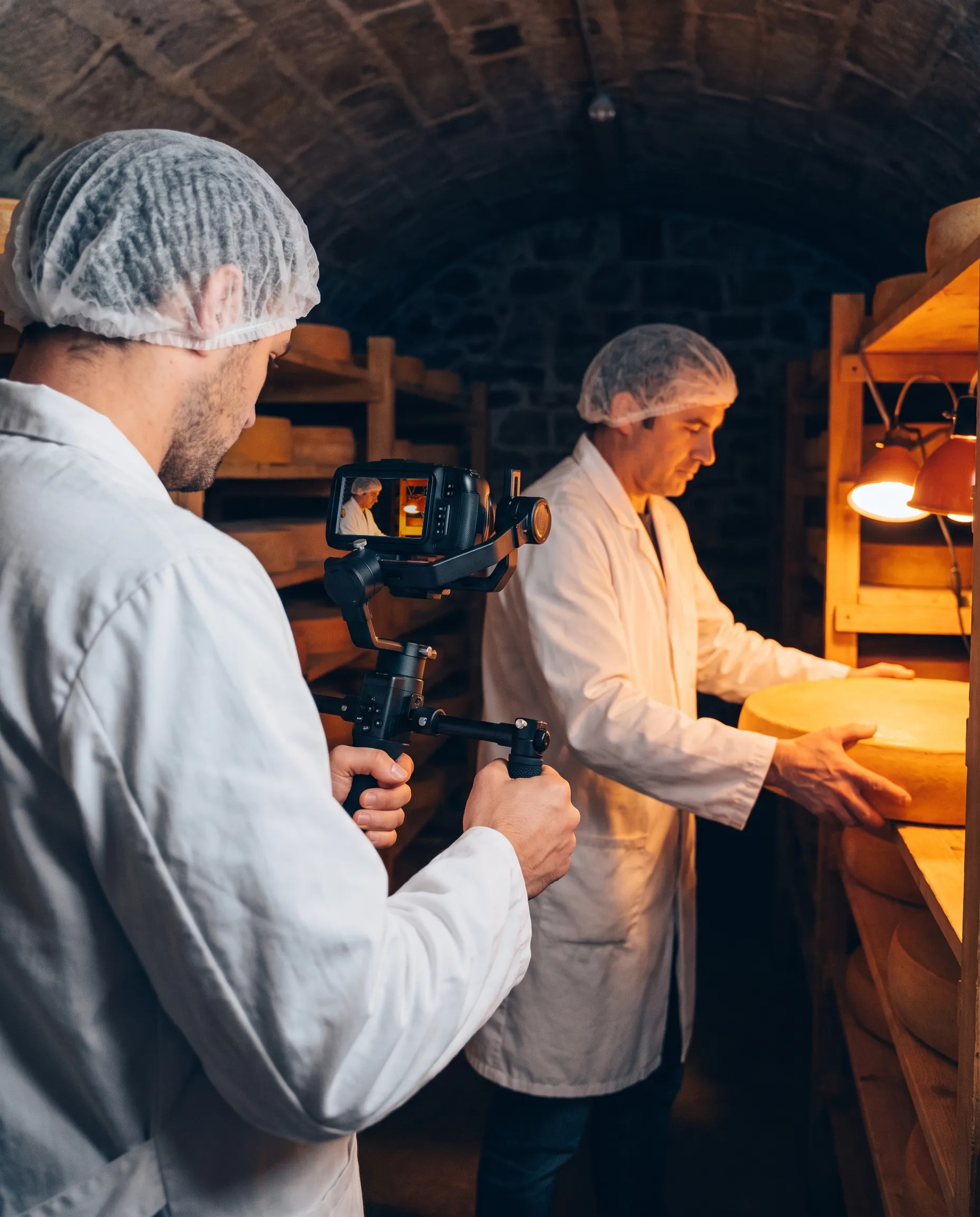 Tournage au stabilisateur dans une cave d’affinage voûtée : un fromager en blouse retourne une meule sur une étagère en bois