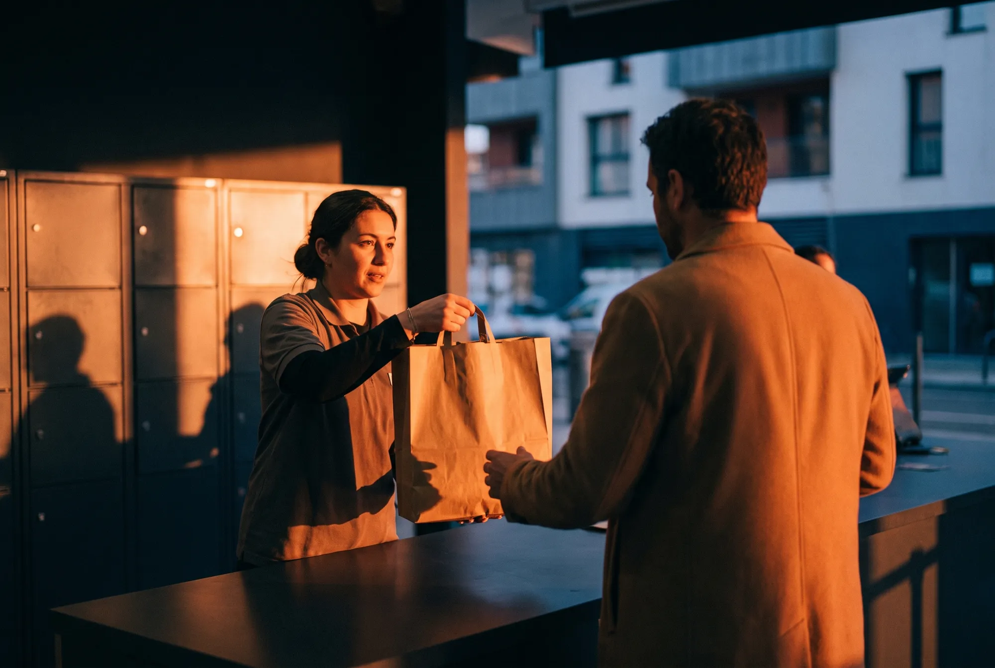Employée tendant un sac en papier kraft à un client au comptoir de retrait des commandes d’un supermarché, casiers en fond