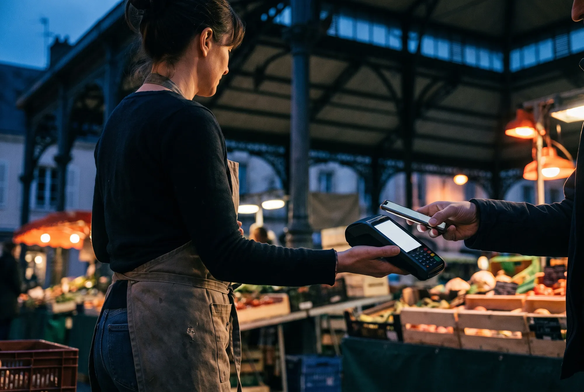 Primeure tendant un terminal de paiement à une cliente qui règle sans contact avec son téléphone sous une halle de marché
