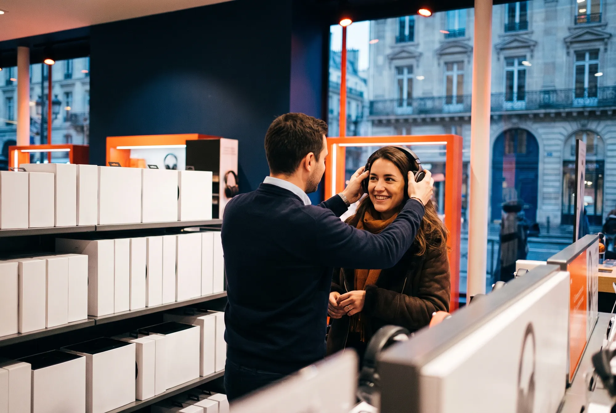 Vendeur faisant essayer un casque audio à une cliente souriante dans le rayon audio d’un grand magasin ouvert sur une rue haussmannienne