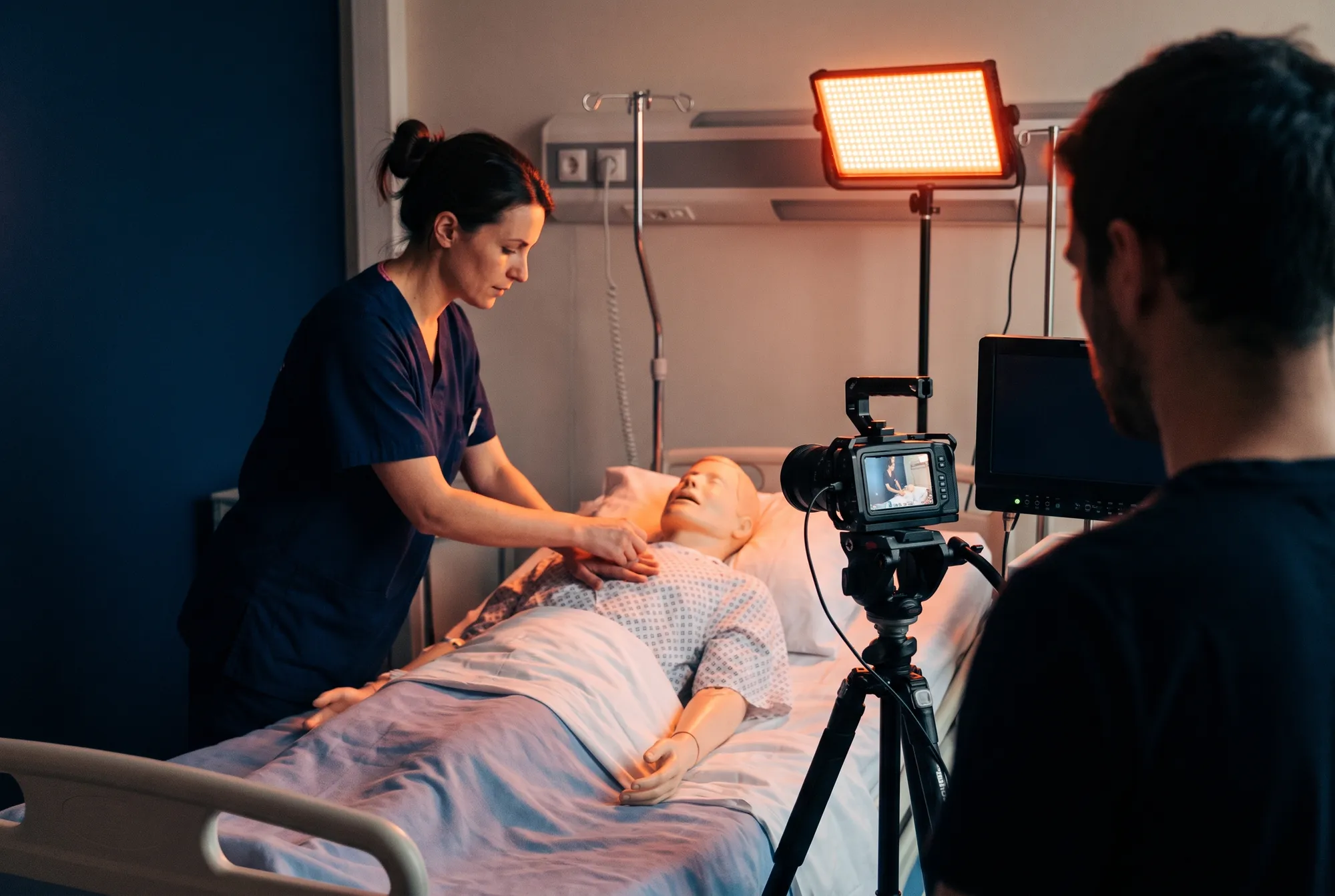 Tournage d'un module de formation en centre de simulation : une infirmière montre un geste sur un mannequin, caméra au premier plan