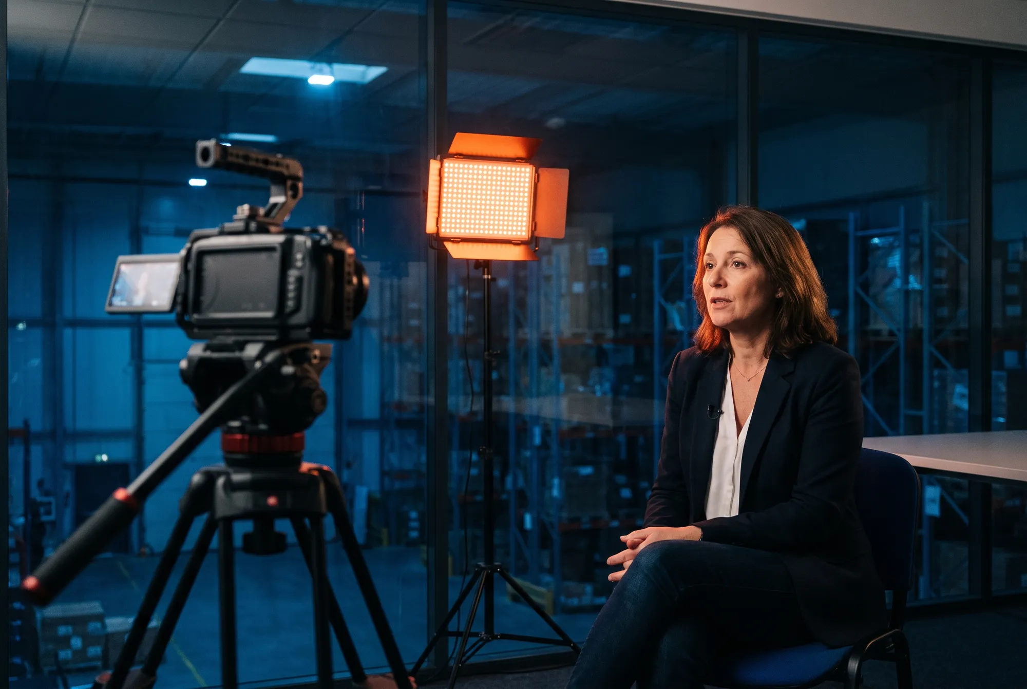 Interview filmée d'une directrice logistique dans son bureau vitré, éclairée par un panneau LED, entrepôt en arrière-plan