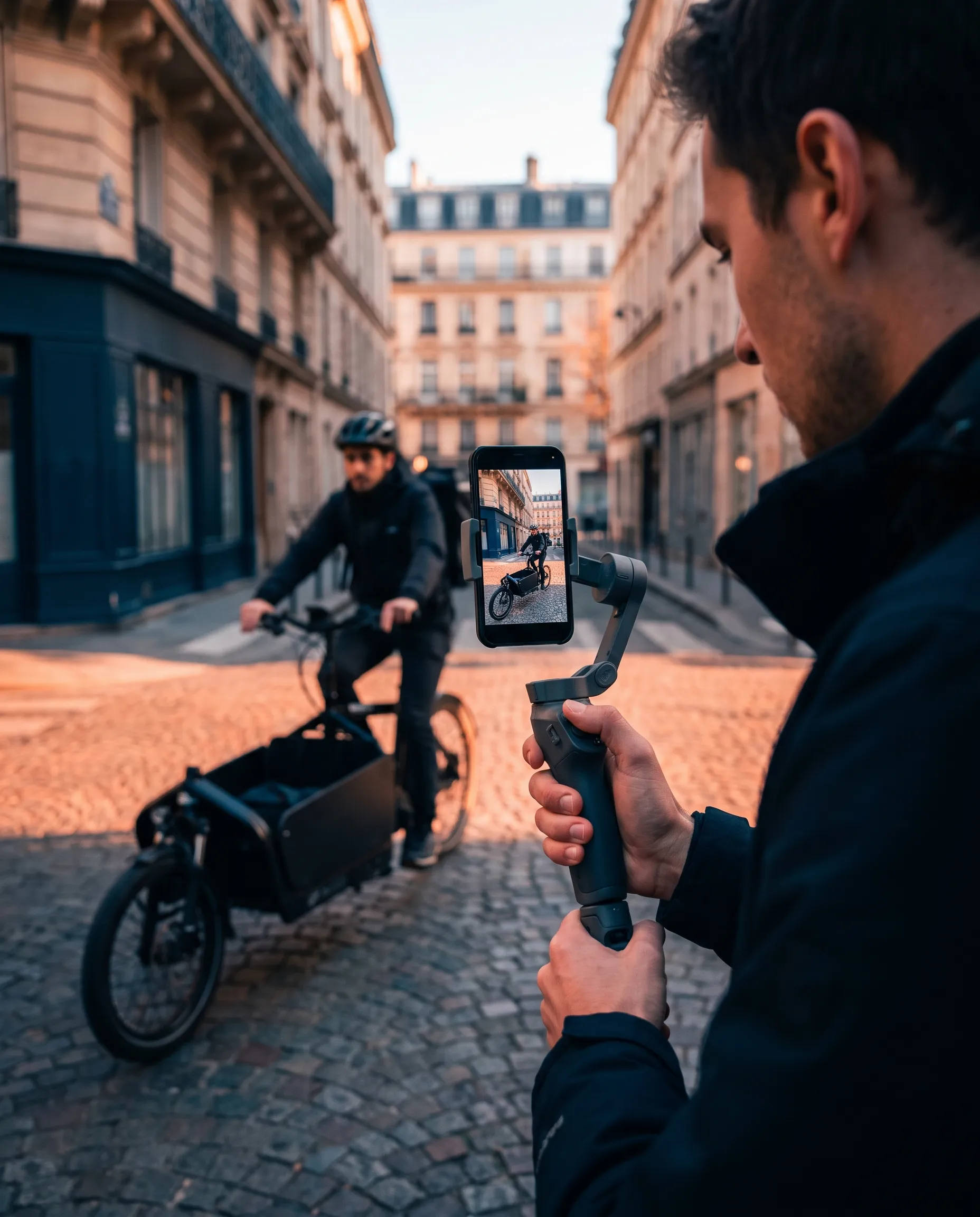 Tournage d'une capsule verticale au smartphone sur stabilisateur tenu à deux mains : un livreur démarre à vélo cargo dans une rue parisienne